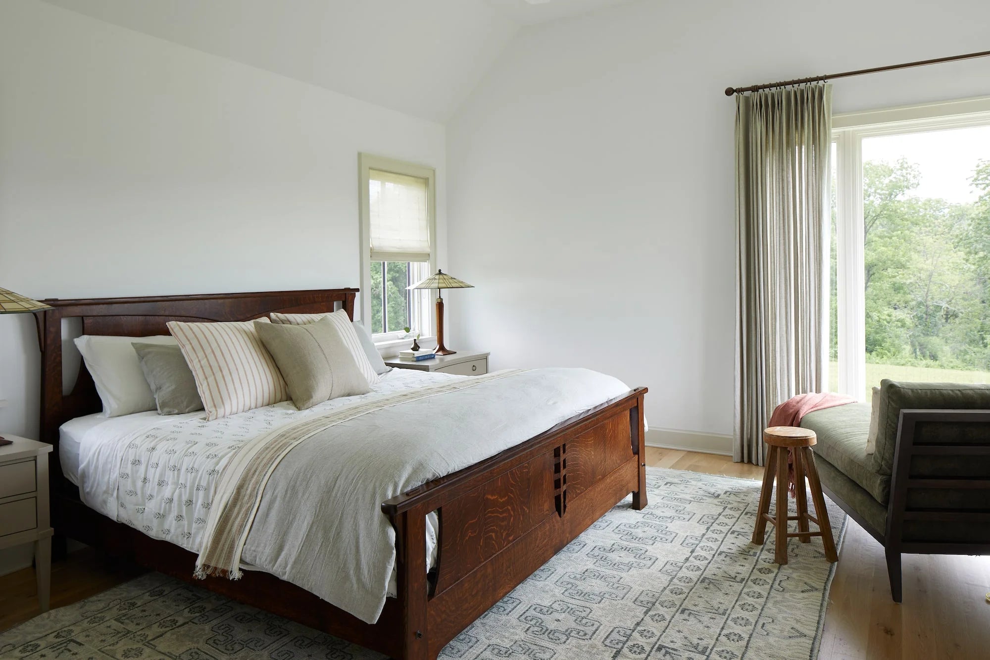 Cold Brook Farm bedroom with dark wood bed frame, layered neutral bedding, soft area rug, and large window with countryside views.