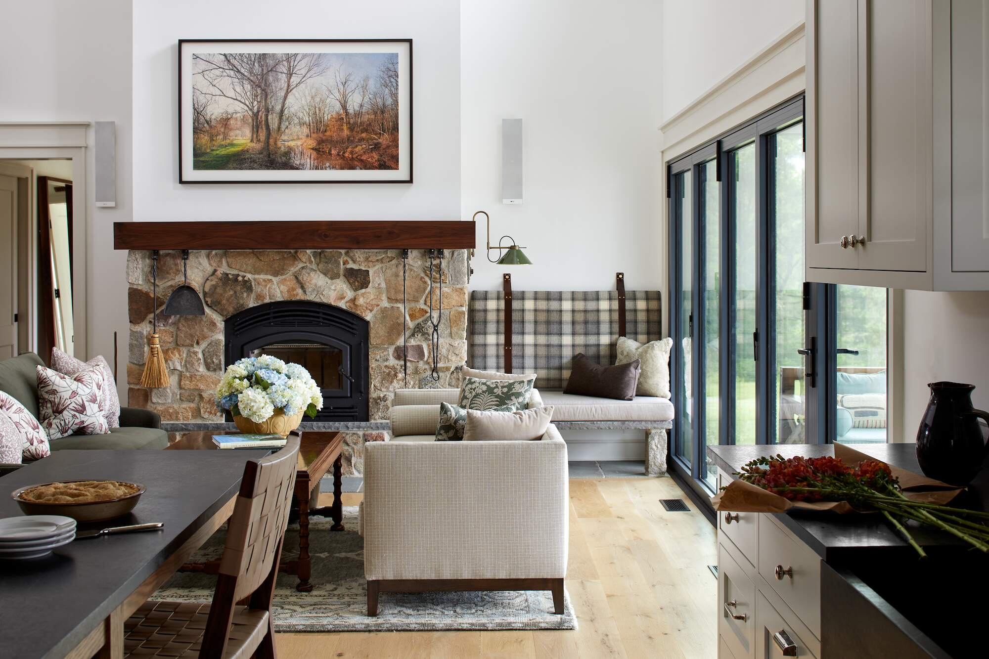 Rustic living room at Cold Brook Farm with stone fireplace, wood mantel, plaid banquette seating, and sliding glass doors to the outdoors.