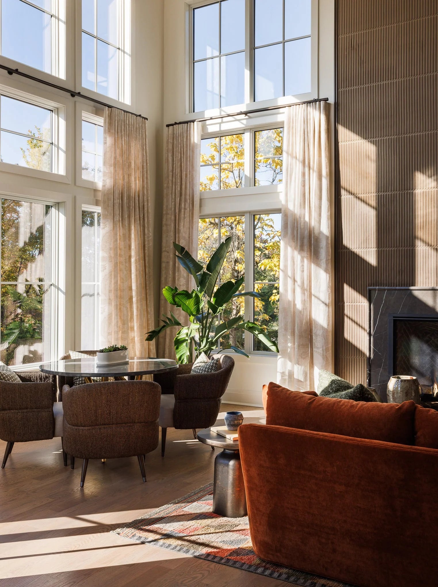 Double-height living room at Twin Brooks with soaring windows, warm neutral furnishings, and a round dining table near a modern fireplace.