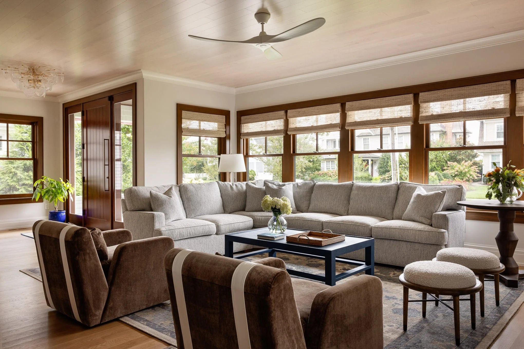 Bright living room with gray sectional sofa, brown velvet chairs, and large windows framed in natural wood.