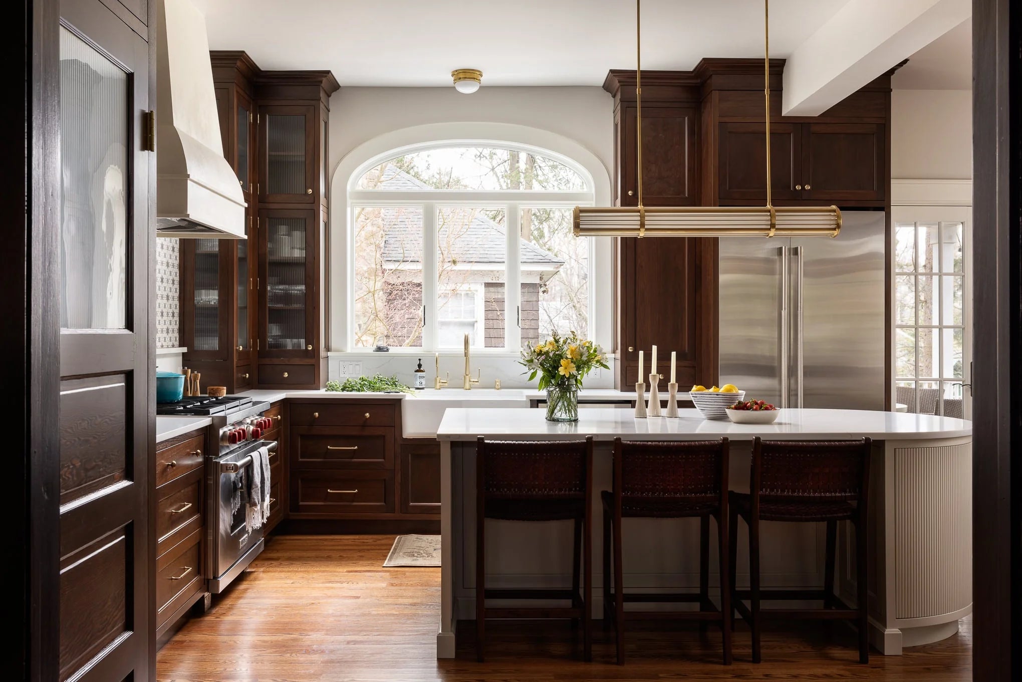 Elegant kitchen at Lawrence featuring rich wood cabinetry, arched window, marble island with brass lighting, and modern chef’s range.