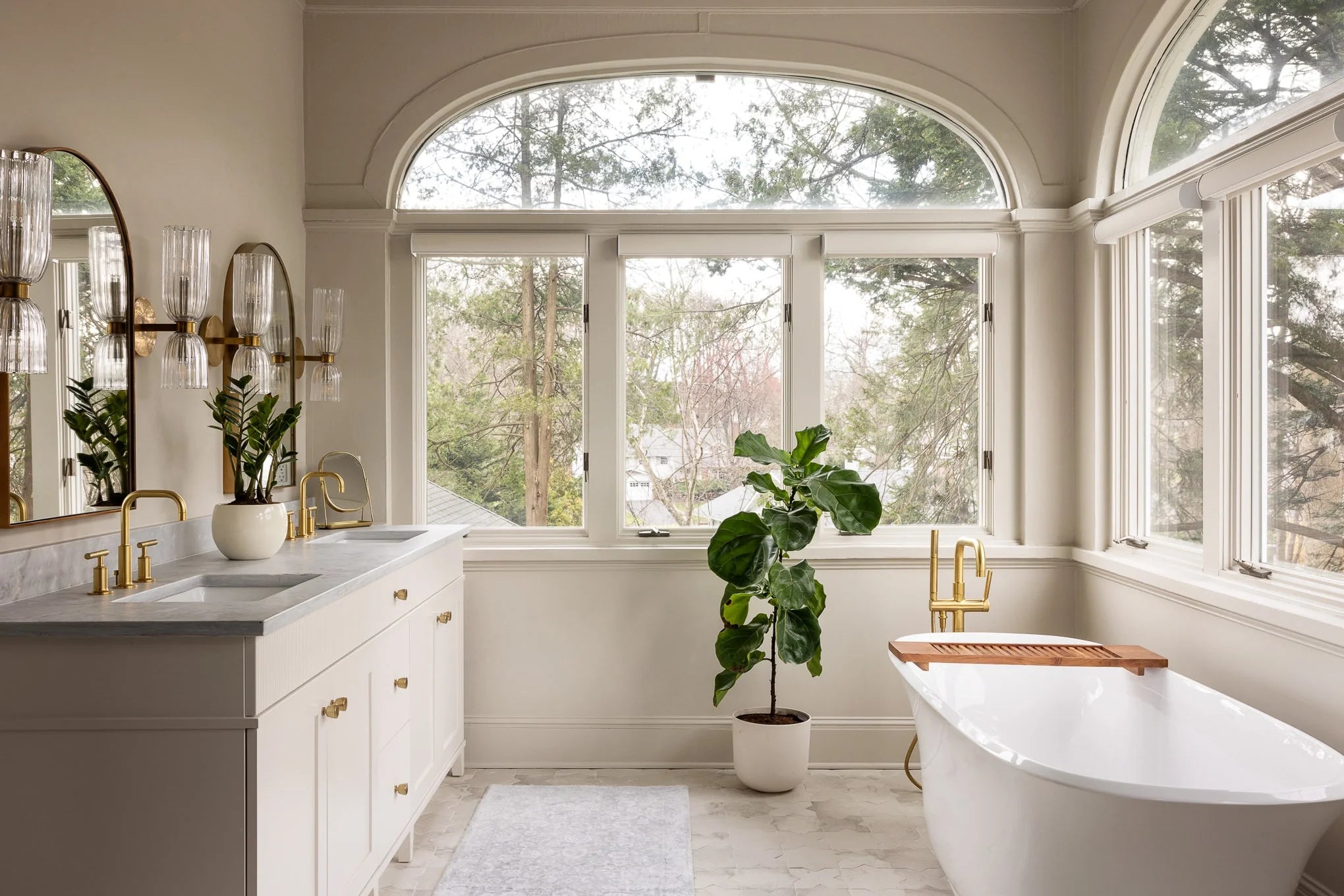 Bright bathroom at Lawrence with freestanding tub, double marble vanity, brass fixtures, and arched windows overlooking the treetops.