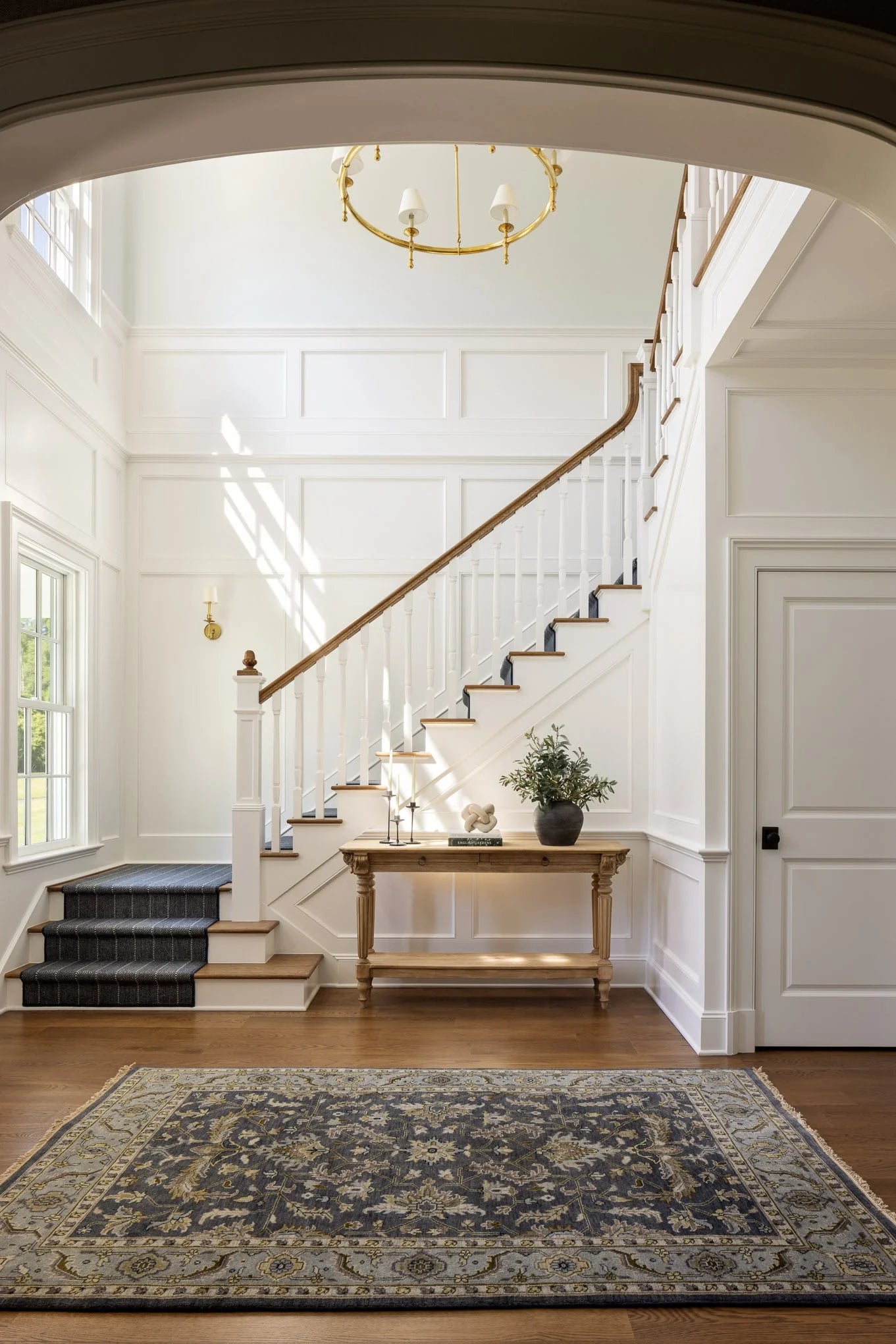 Bright entry foyer with grand staircase, paneled white walls, gold chandelier, and patterned rug at Moonlight project.
