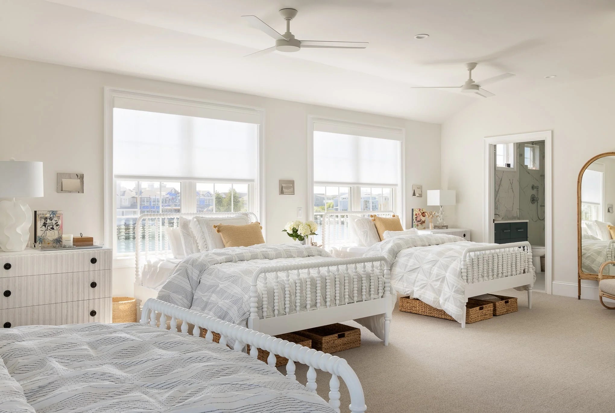 Bright coastal bedroom with three white spindle beds, light gray bedding, and woven baskets for storage beneath, overlooking waterfront views.