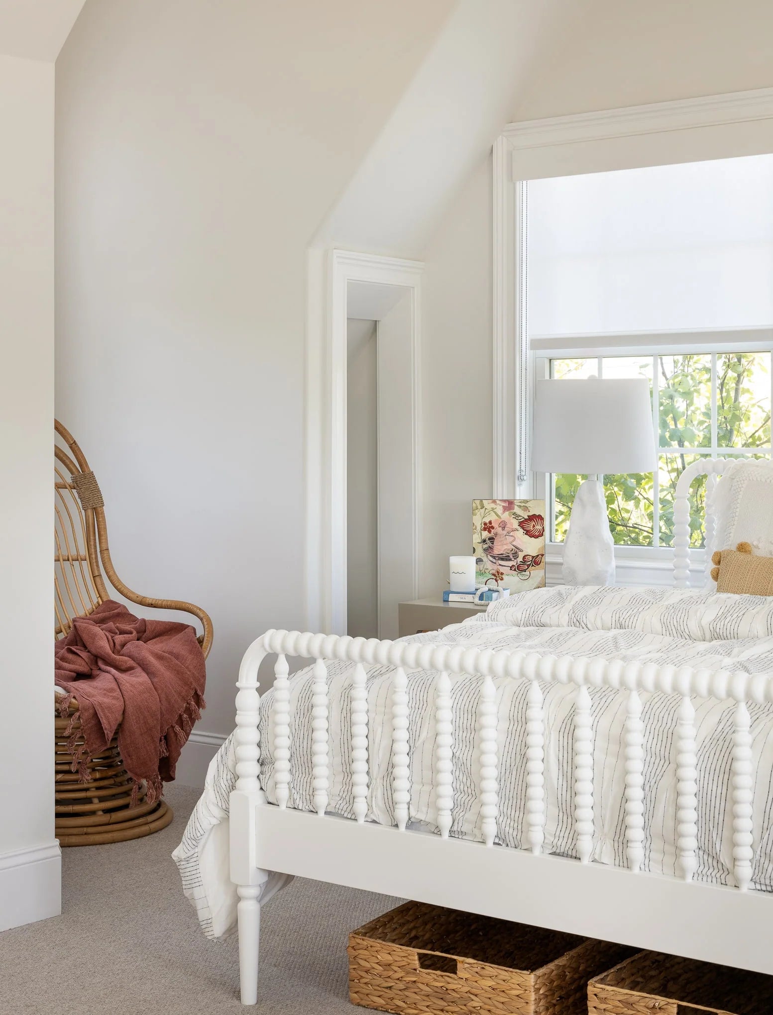 Corner of a serene bedroom with a white spindle bed, rattan chair draped with a terracotta throw, and soft natural light through the window.