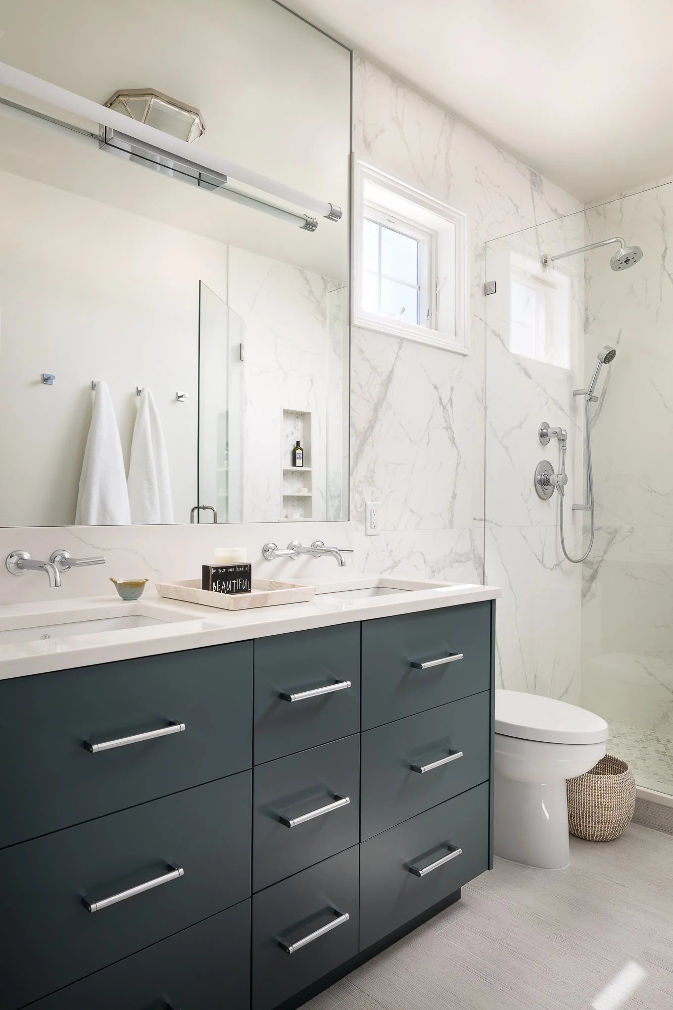 A sleek bathroom featuring a double vanity with navy drawers, chrome hardware, marble shower walls, and a large mirror above the sinks.
