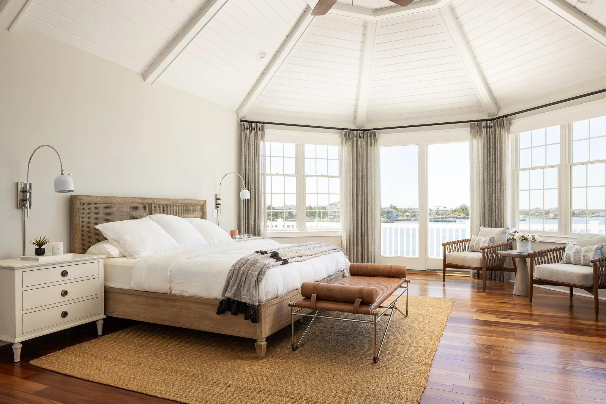 A bright coastal bedroom with a vaulted white ceiling, wood bed with rattan headboard, layered neutral bedding, and leather bench at the foot. Large windows with gray drapery overlook the water.
