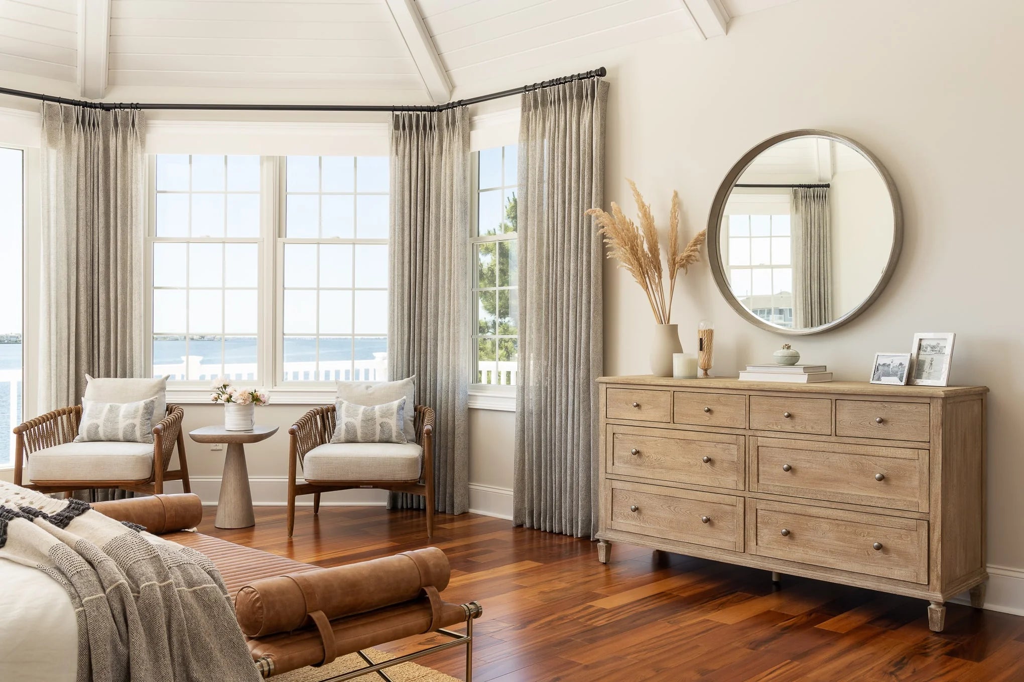 A natural wood dresser with a round mirror above, styled with pampas grass, framed photos, and books, sits across from seating by the windows.