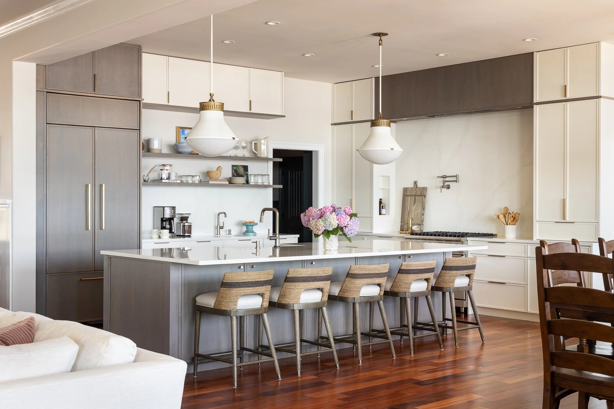Large kitchen island with woven bar stools, modern pendant lights, and hydrangea centerpiece, overlooking dining area – Haven Bay design.