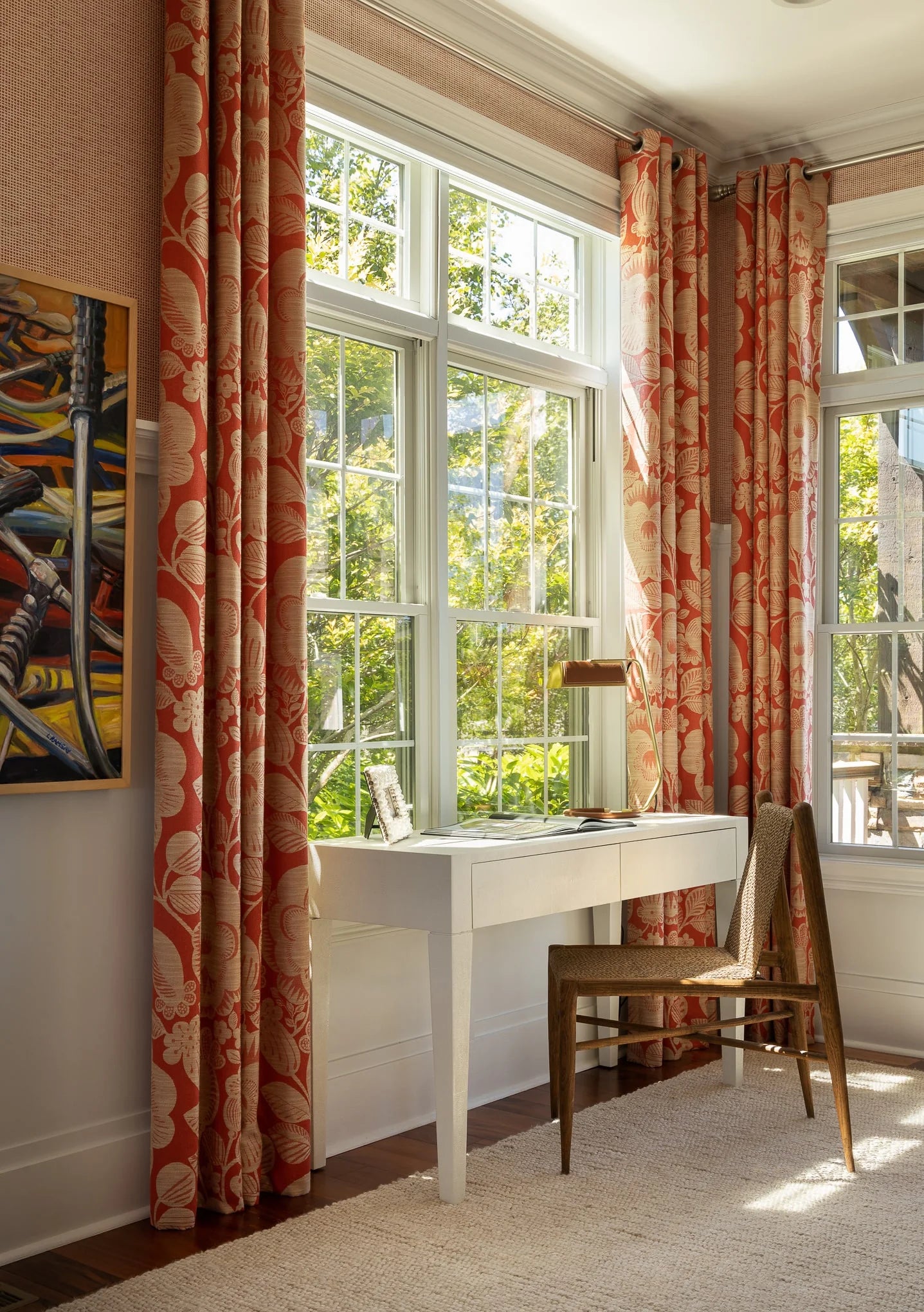 Bright home office corner with a white writing desk and wooden chair, framed by bold red and cream floral curtains and large windows bringing in natural light.