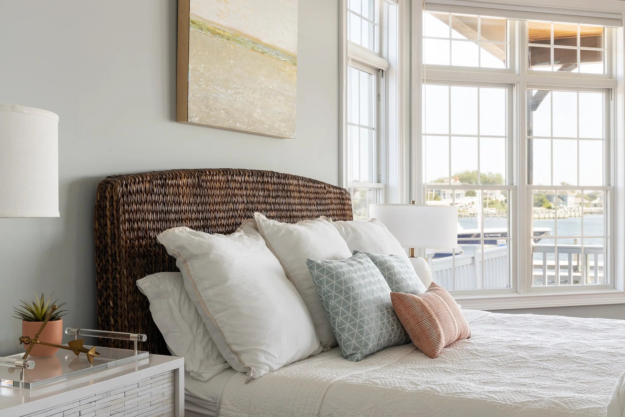 Coastal bedroom with a woven rattan headboard, crisp white bedding, and accent pillows in soft blue and blush tones, set beside large windows overlooking the water.