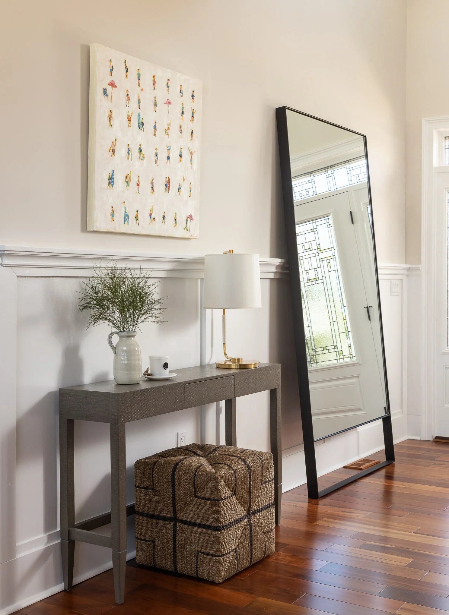 Entryway console with ceramic jug, lamp, woven pouf, and leaning floor mirror against white wainscoting.
