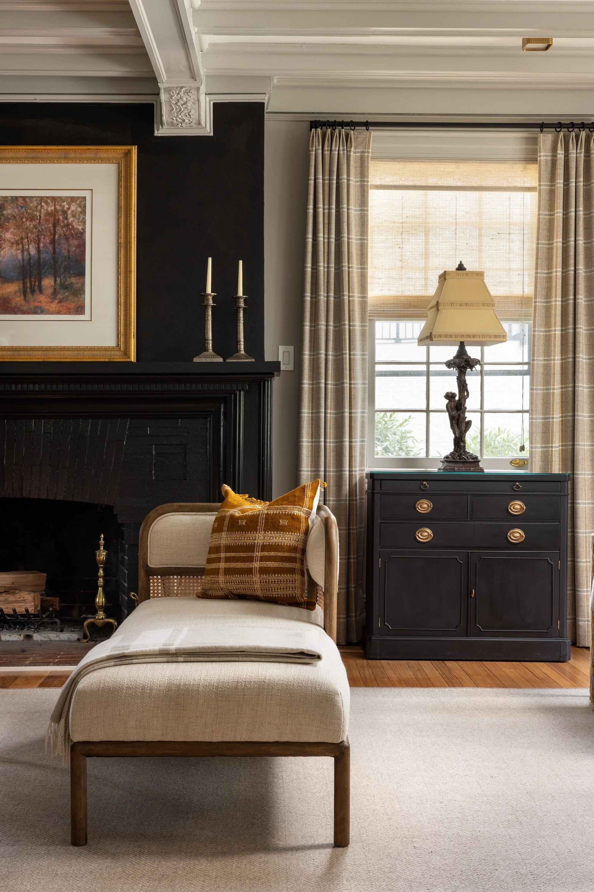 Detail of Stoneleigh living room by AHD&Co with cane daybed, mustard accent pillow, black fireplace, and tailored neutral window treatments.