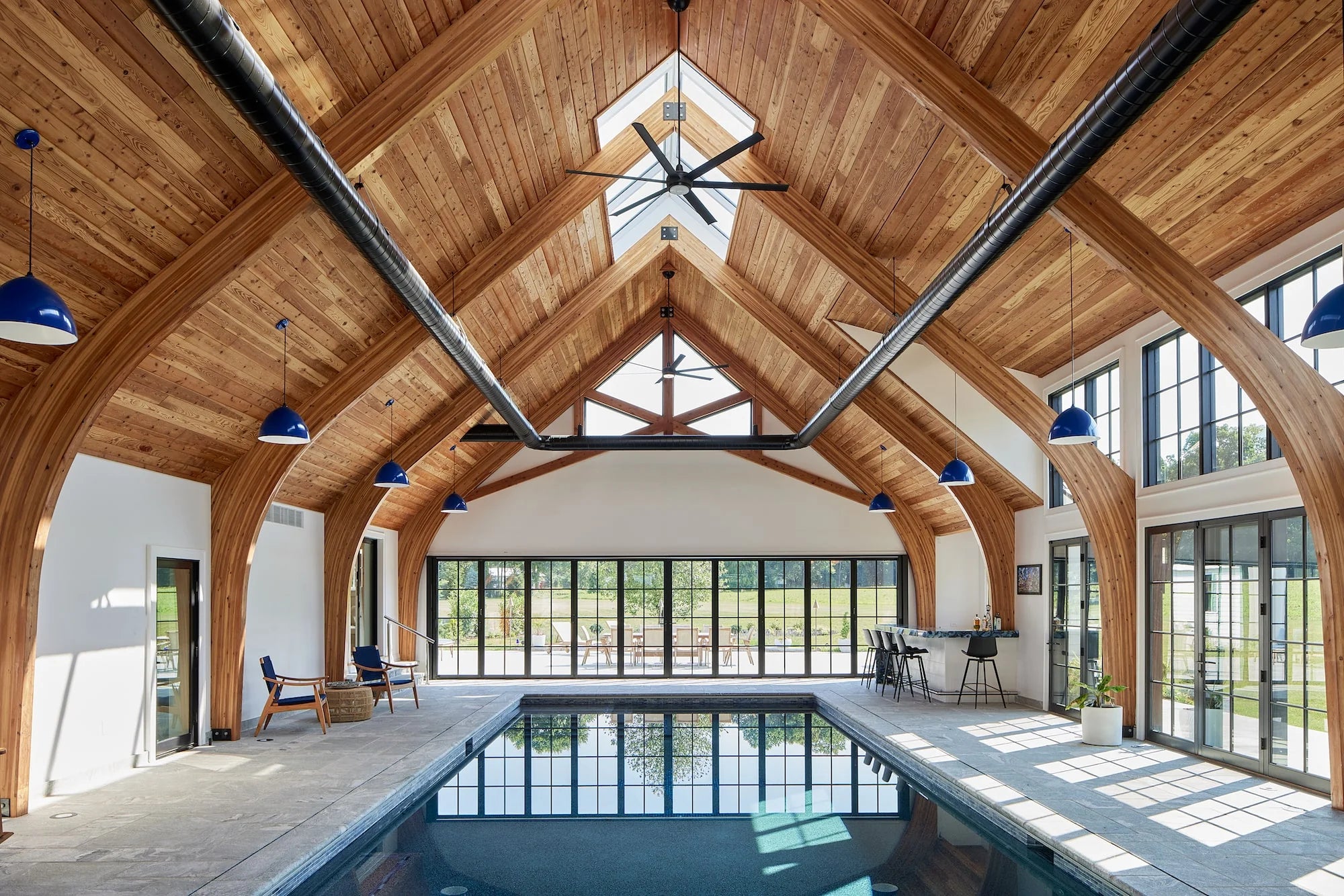 Vaulted indoor pool room with exposed wood beams, blue pendant lights, and floor-to-ceiling windows at Airport Road