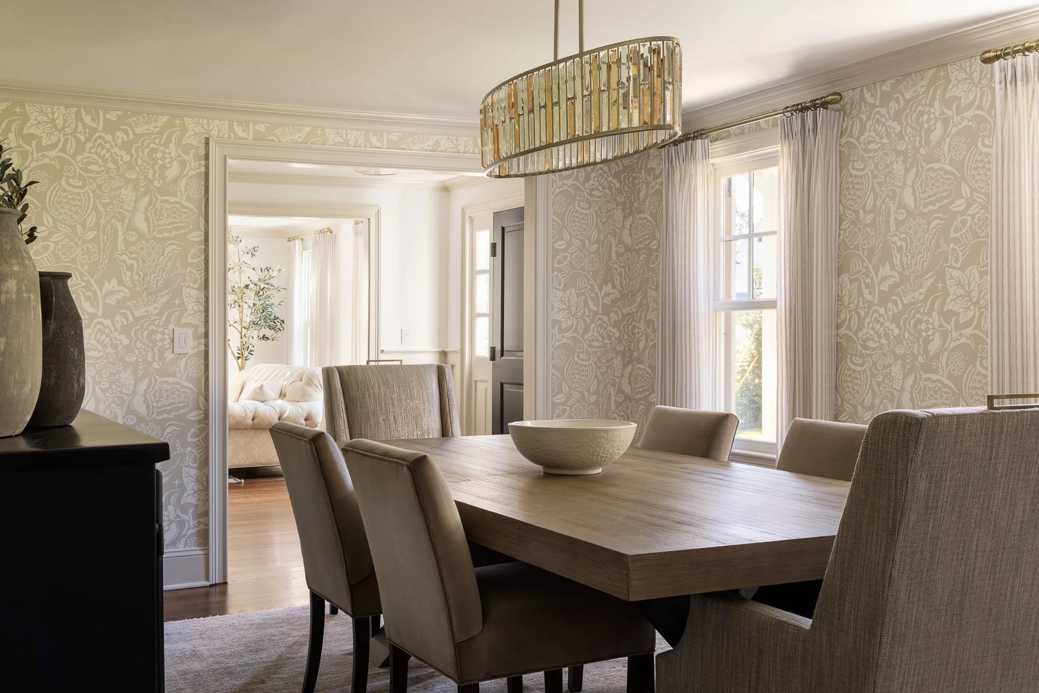 Neutral dining room with patterned wallpaper, upholstered chairs, and a modern gold chandelier above a wood table.