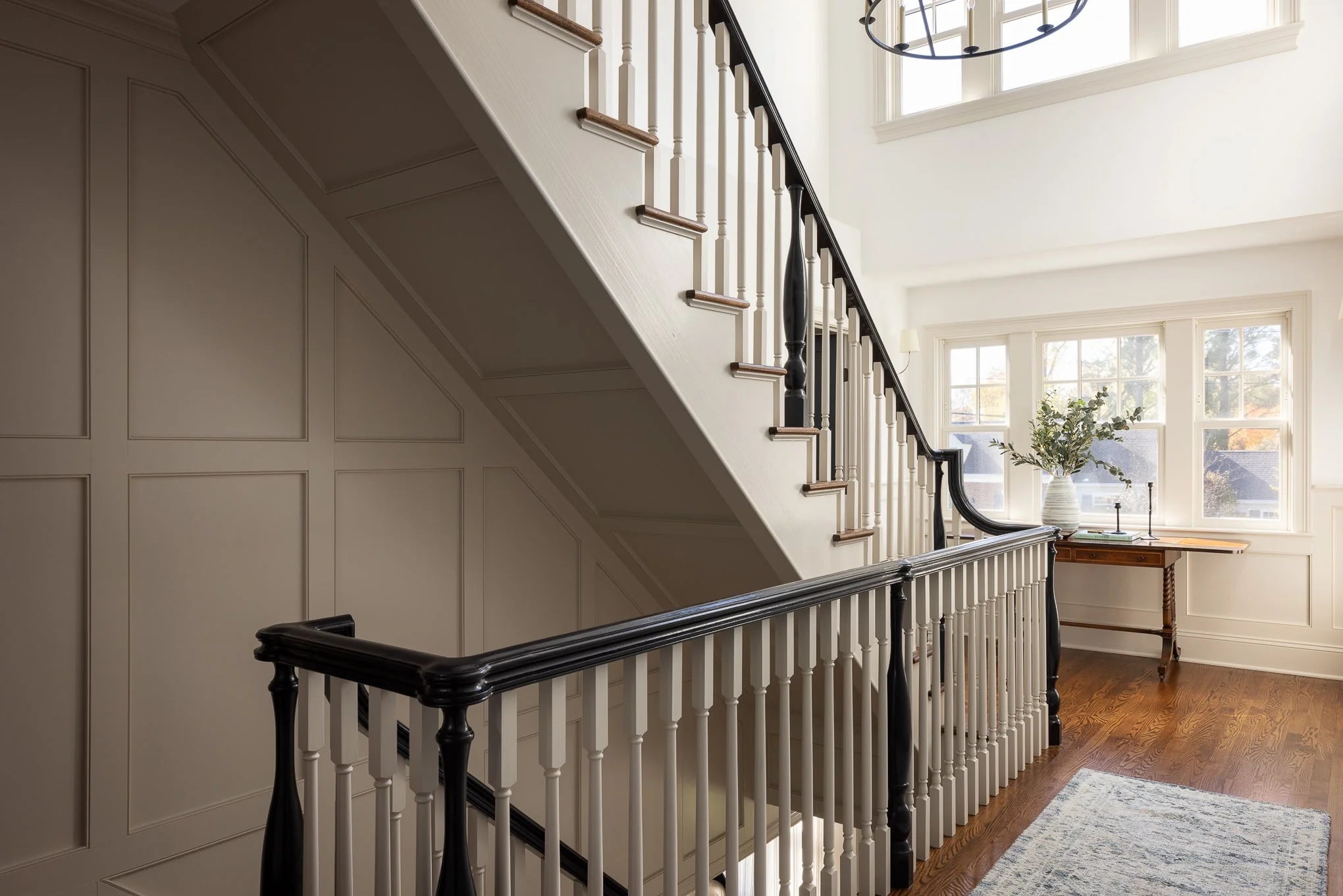 Elegant staircase with white spindles and dark railing, paneled wall detailing, and natural light from upper windows.