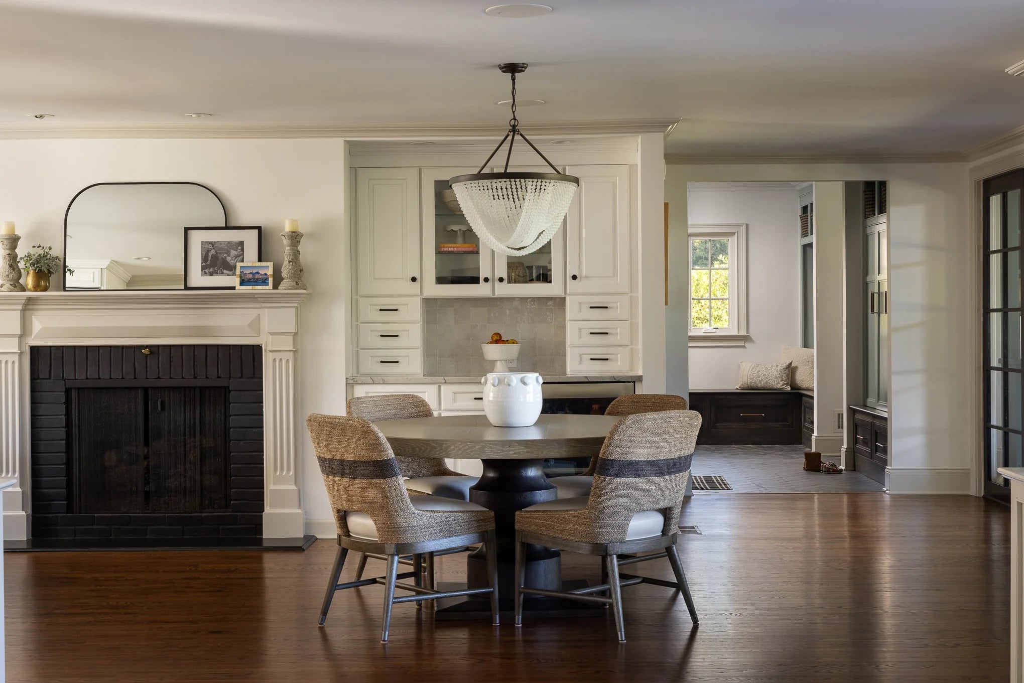 Transitional dining space with a round pedestal table, woven chairs, and a black fireplace accented by a large mirror and white cabinetry.