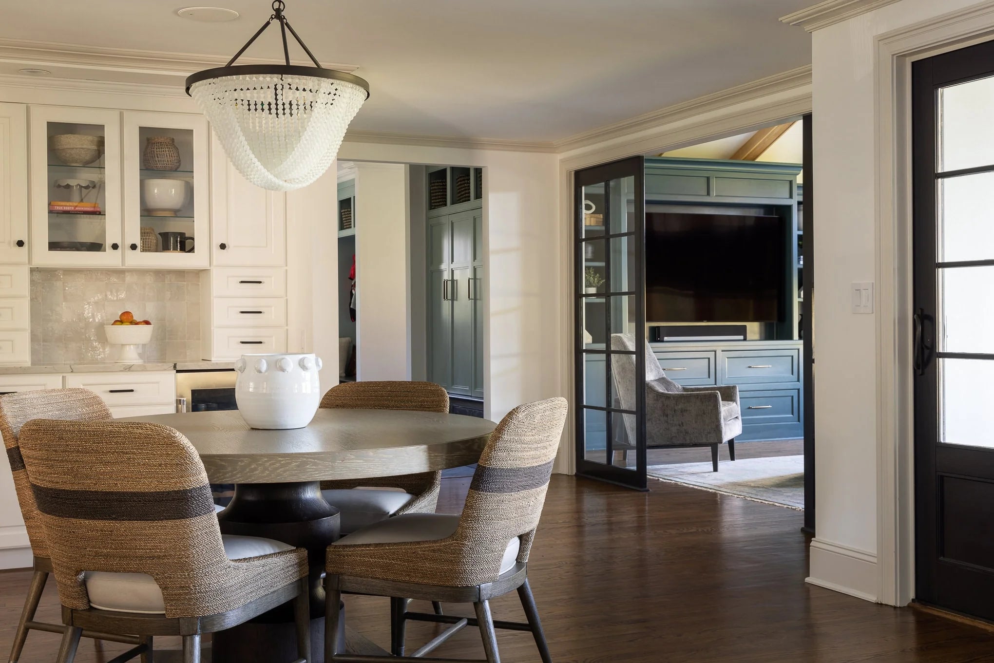 Neutral dining nook with round wood table, woven chairs, and a beaded chandelier, opening into a media room with built-in cabinetry and French doors.